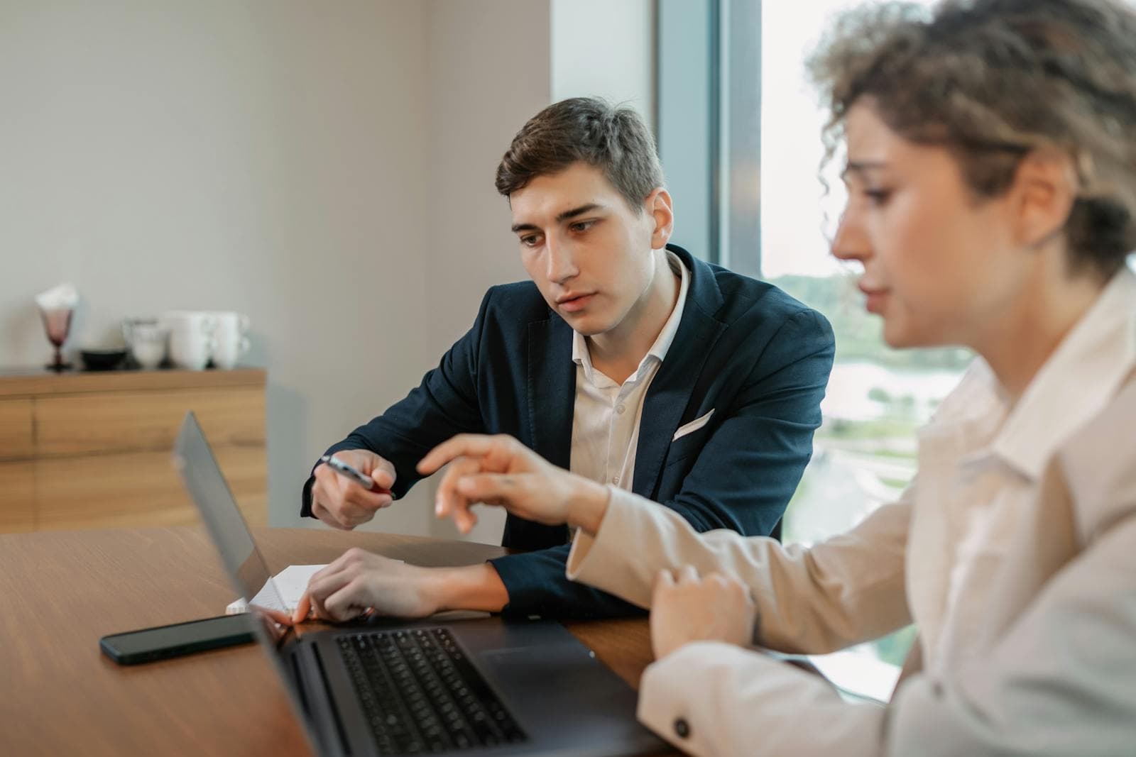 Two professionals reviewing a workflow on a laptop together at a conference table, one in a navy blazer pointing at the screen while explaining the system to a colleague taking notes, the embedded delivery model that built Palantir's enterprise franchise