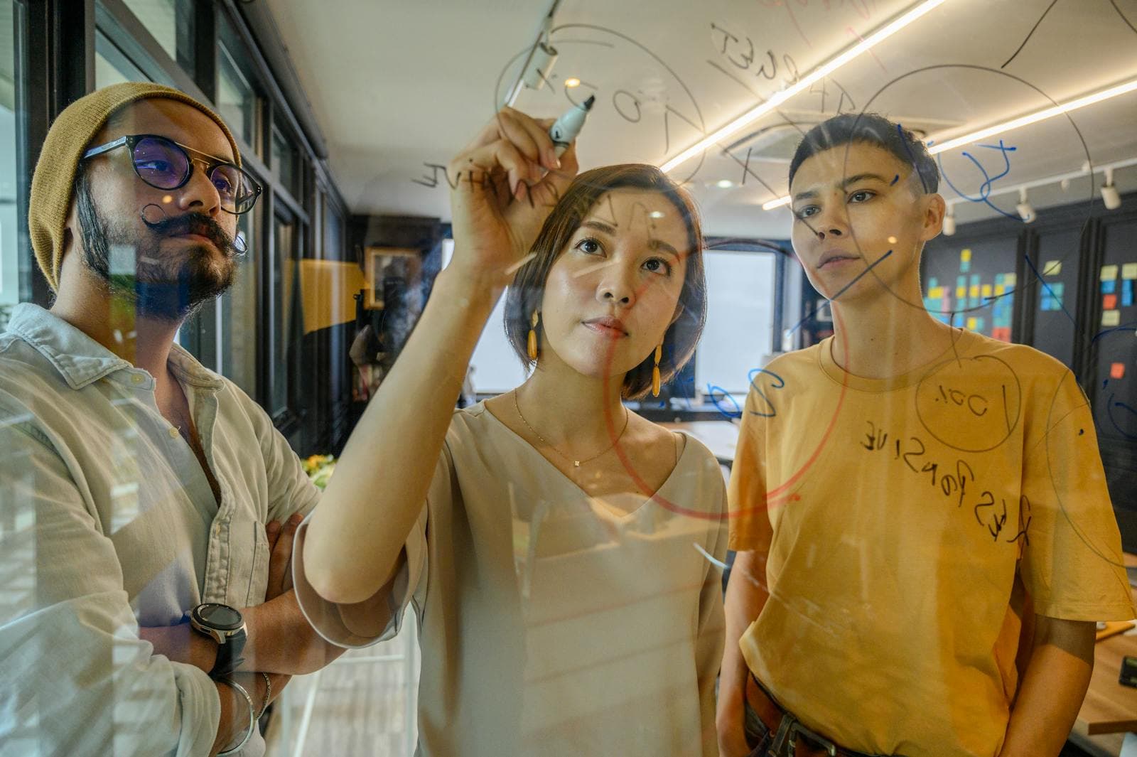 A woman in a black sleeveless top sketching system diagrams on a glass whiteboard during a planning session, the architecture conversation every team should have before the first line of production code ships